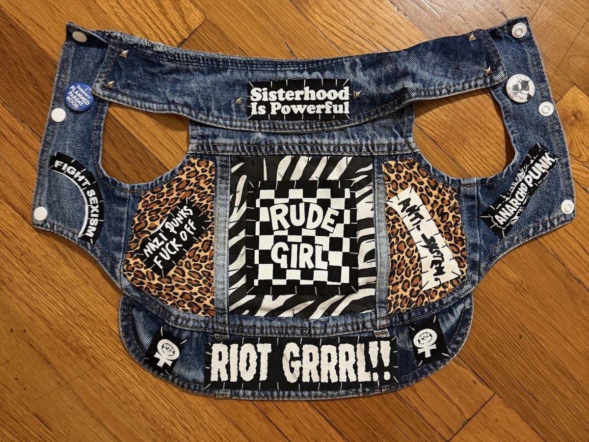 The Rude Girl vest laid flat: a checkerboard Rude Girl back piece on zebra print, leopard-print side panels with Nazi Punks Fuck Off and Anti-System strips, Sisterhood Is Powerful at the collar, Fight Sexism and Anarcho Punk patches, a chenille RIOT GRRRL!! strip across the hem, gold studs and pins